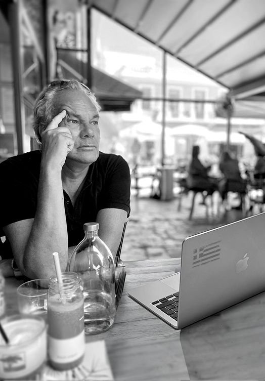 Michael Saupe, Porträt in einem Café auf Kreta, nachdenklich am MacBook mit griechischer Flagge